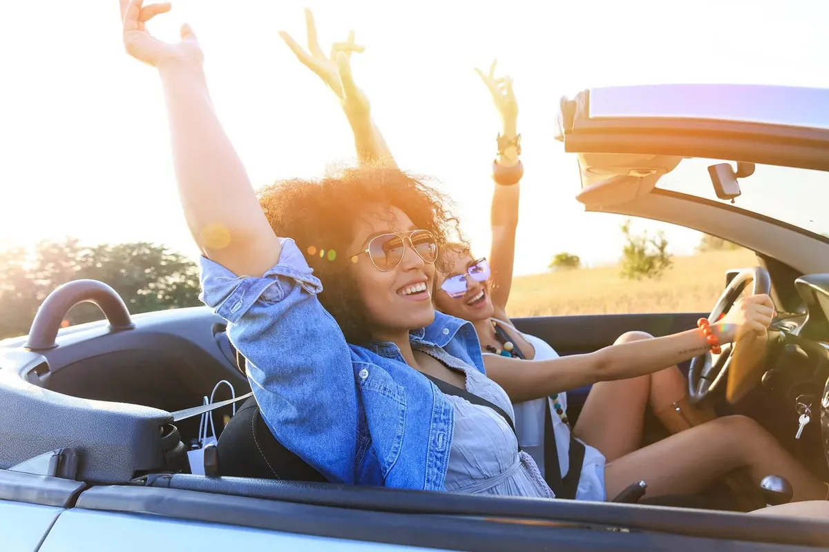 Zwei Frauen fahren lachend mit erhobenen Armen in einem offenen Cabrio